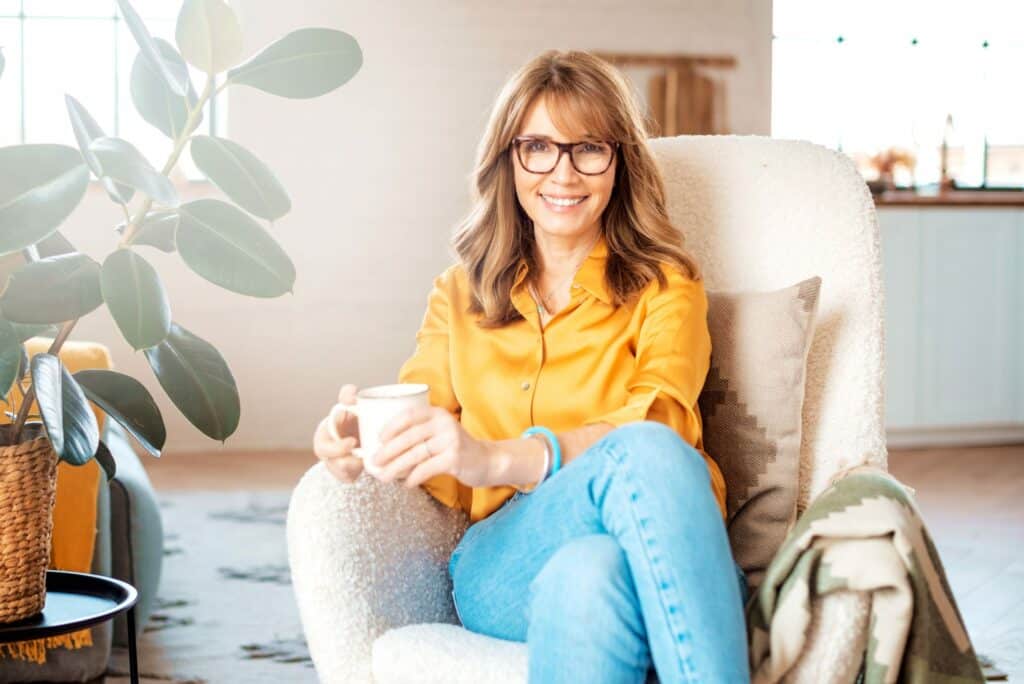 A female doctor, thriving general practitioner with the freedom of consulting, smiles contentedly. She relaxes in a comfortable chair, holding a mug, with a large plant and a bright interior scene in the background. Her casual attire - glasses, long hair, yellow blouse, and jeans - reflects her newfound flexibility in her consulting career.